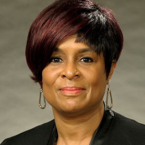 A professional headshot of Stephanae's smashing asymmetrical hairstyle with burgundy highlights. The muted Coral Cutie lipstick topped with a peach colored gloss provides a nice contrast against the gray backdrop. She is wearing a black dress and black tuxedo jacket trimmed in faux leather, silver statement necklace, and silver drop earrings.